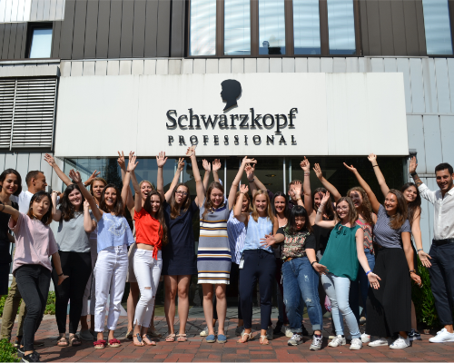 Et mangfoldig Henkel-team som står og heier foran Schwarzkopfs profesjonelle bygning og løfter armene 