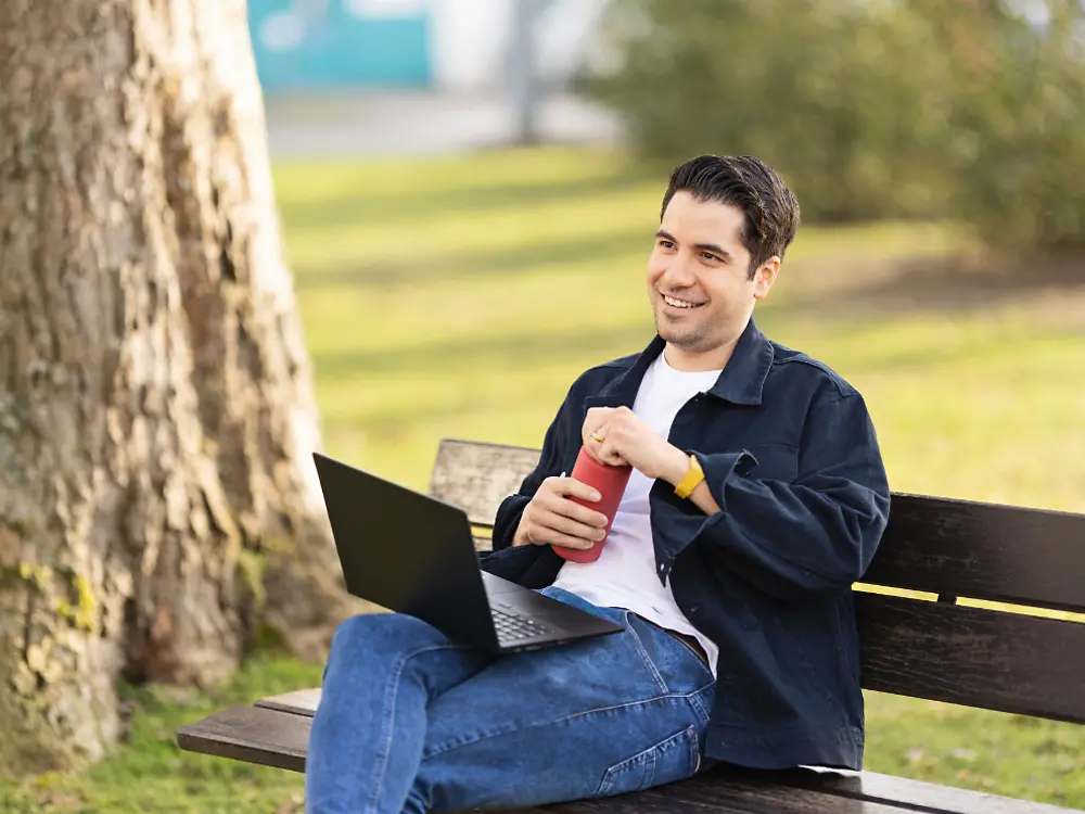 En Henkel-ansatt sitter på en benk i en park og jobber på den bærbare datamaskinen sin. 