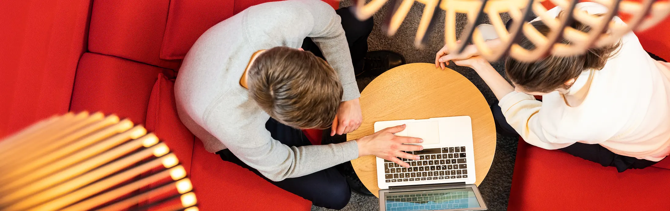 To personer sitter på en rød sofa og jobber sammen på en datamaskin.