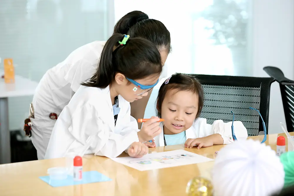 To barn og en kvinne i en forskerfrakk farger et bilde ved et bord
