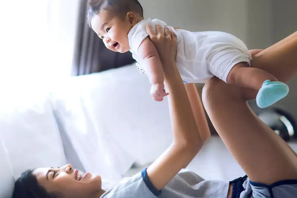 Mor løfter og leker med en smilende baby innendørs i et lyst hjemmemiljø.