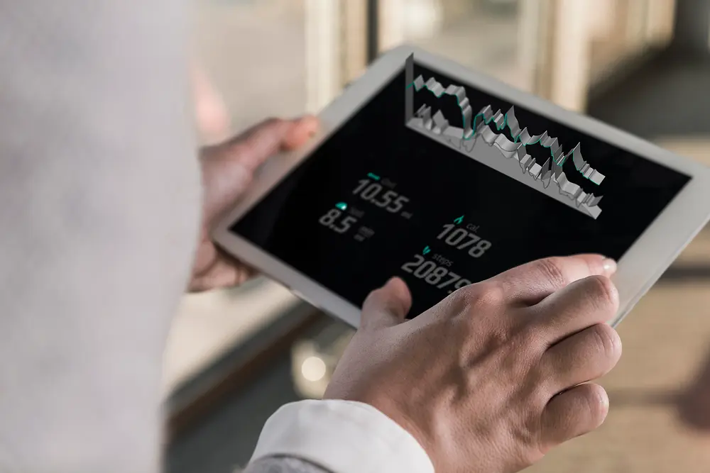En person som holder et nettbrett med digitale kart og tallopplysninger, sett over skulderen i et lyst rom.